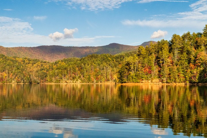 Serene lake with tree-covered hills reflecting on calm water under a partly cloudy sky.