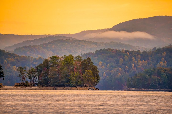 Island with trees on a lake, mountains in background, under a warm yellow sky.