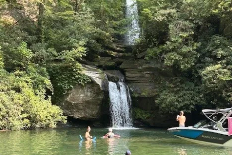 People swimming near a waterfall in a forested area with a boat nearby.