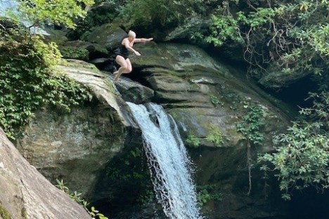 Person jumping off a rock beside a small waterfall into a forest pool.