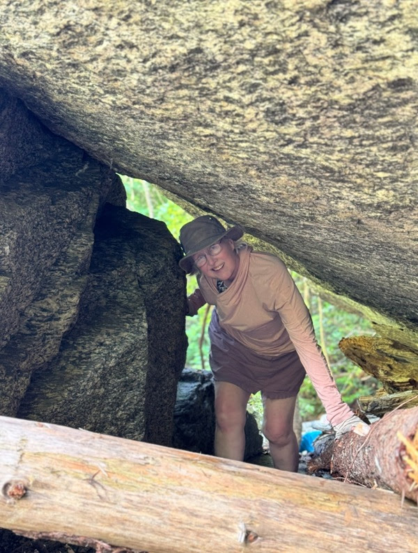 Person in outdoor attire crouching under large rocks in a wooded area.
