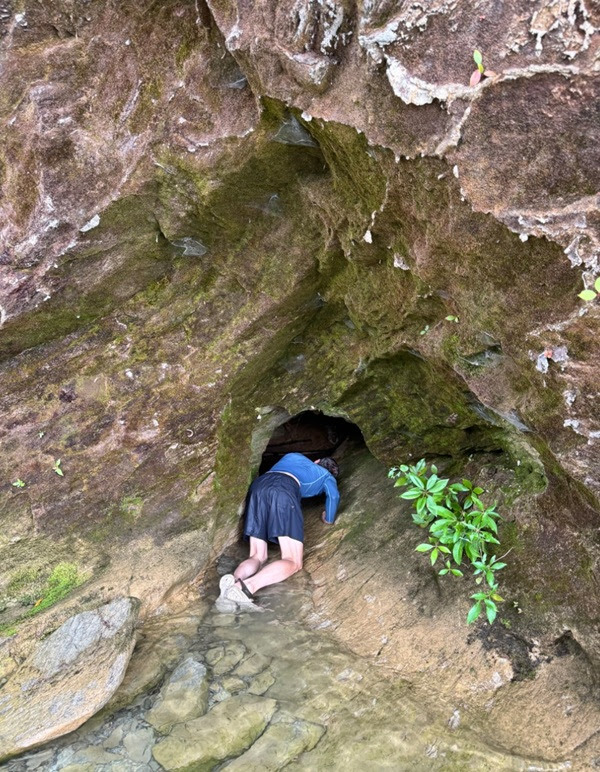 Person in blue jacket crawling into a small cave entrance on a rocky surface.