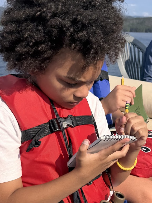 Child in red life jacket writing in notebook on a boat.