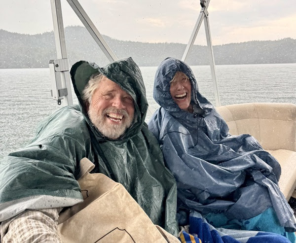 Two people in rain ponchos smiling on a boat with mountains and water in the background.
