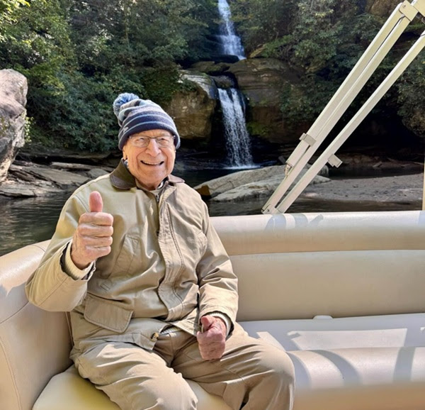 Elderly man in warm clothes smiling and giving thumbs up on a boat with a waterfall in the background.