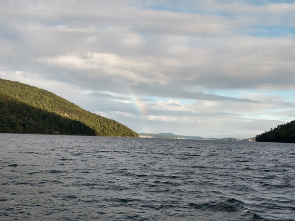 Lake view with a hill and a faint rainbow in a cloudy sky.
