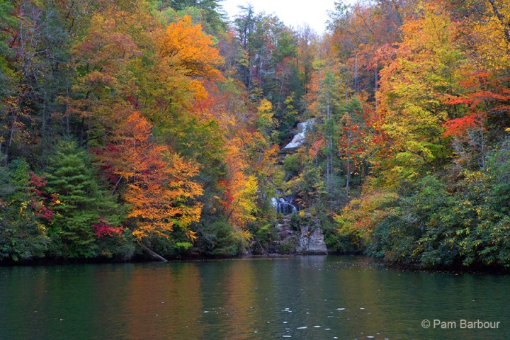 Waterfall amidst colorful autumn trees by a tranquil lake.