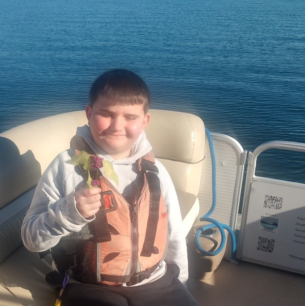 Child on a boat holding a small bouquet, wearing a life jacket and smiling, with water in the background.