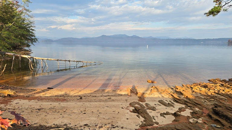 Sandy lakeside with driftwood and rocky shore, overlooking calm water and distant mountains under a cloudy sky.