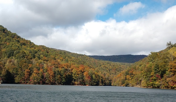 Lake surrounded by forested hills with autumn foliage under a cloudy sky.