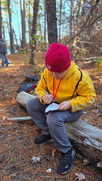 Child in a red hat and yellow jacket writing while sitting on a log in a forest.