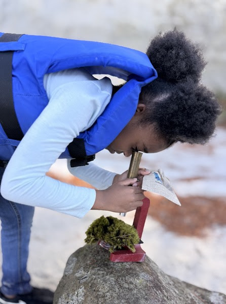 Child examining moss with magnifying glass on a rock, wearing a blue vest.
