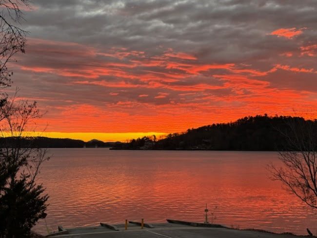 Dramatic red-orange sunset over a lake with silhouetted trees and a cloudy sky.