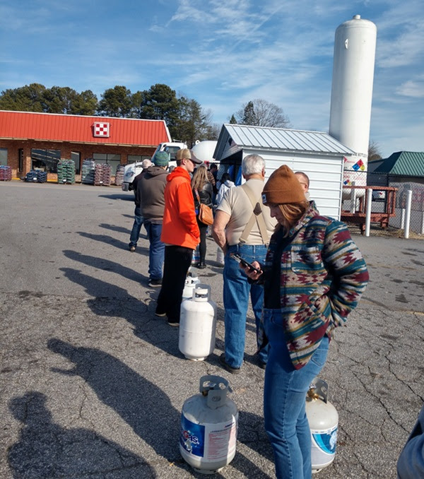 People standing in line with propane tanks outside a store on a sunny day.
