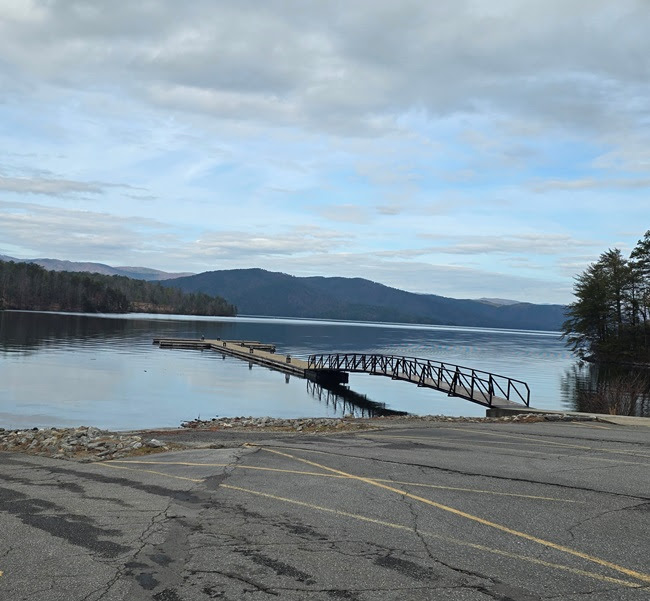 Lakeside dock extending into calm water with mountains and cloudy sky in background.