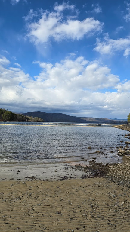 Sandy shoreline with a river, mountains in background, and partly cloudy sky.