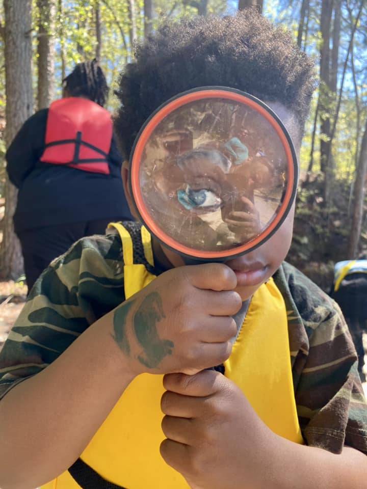 Child holding magnifying glass to eye, wearing camo shirt and yellow vest.