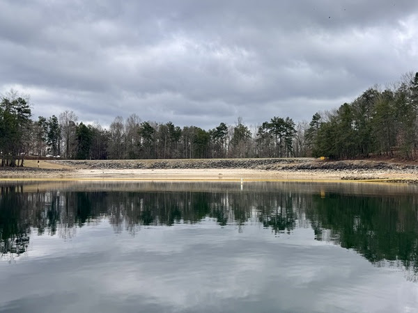 Lake with trees and rocky shoreline under cloudy sky, reflected in water.
