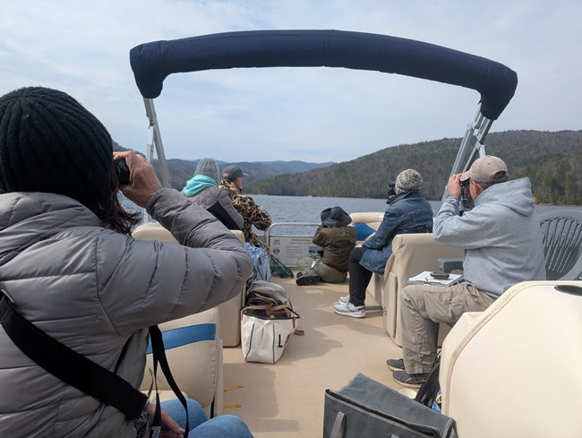 People on a boat using binoculars to view distant mountains and a lake.