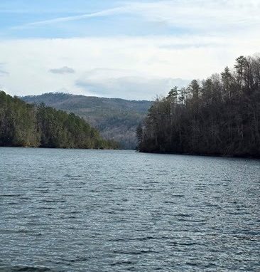 Lake with forested shoreline and distant hills under a partly cloudy sky.