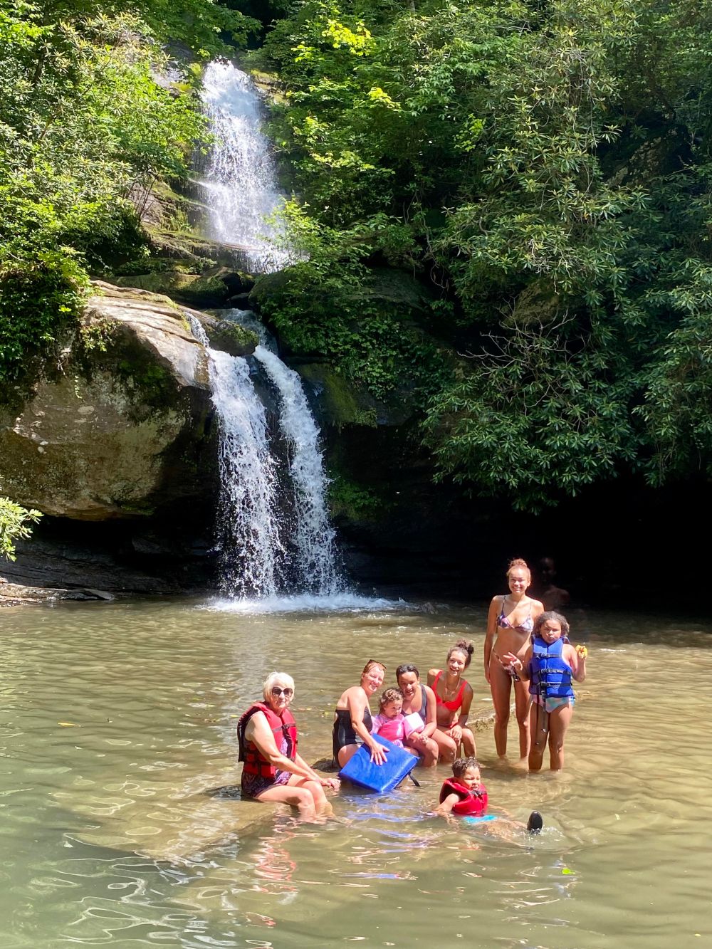 Group of people in a pool below a waterfall surrounded by greenery.