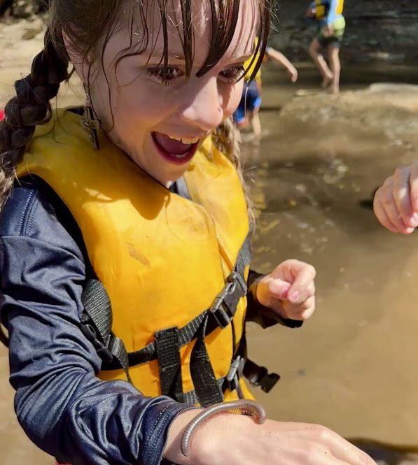 Girl with a yellow vest and wet hair holds a small worm outdoors.