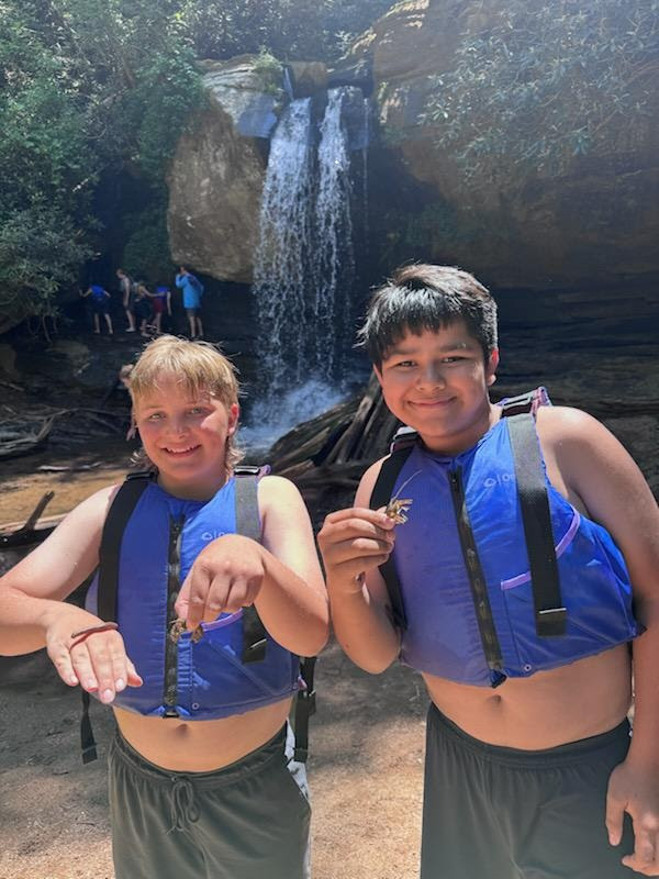 Two boys in blue life jackets holding small items in front of a waterfall.