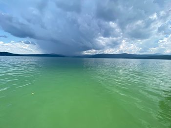 Calm lake with greenish water under a cloudy sky, distant hills in the background.