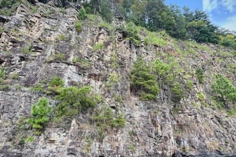 Rocky cliff with trees above, next to blue water under a partly cloudy sky.