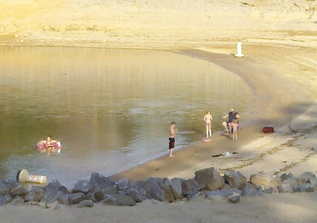 People enjoying a sunny day by a lake with sandy shoreline and rock border.