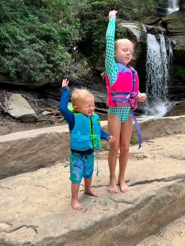 Two children in life vests stand on rocks by a waterfall, one with eyes shut and the other with a raised hand.