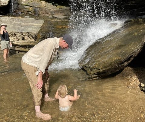 Man and toddler near a waterfall in a rocky area.