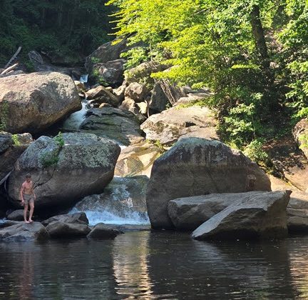 Person standing near boulders by a small waterfall and forested area.