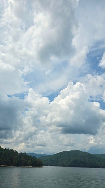 Lake with tree-covered hills under a sky filled with fluffy clouds.