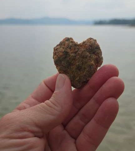 Hand holding a heart-shaped rock by a calm lake with distant hills.