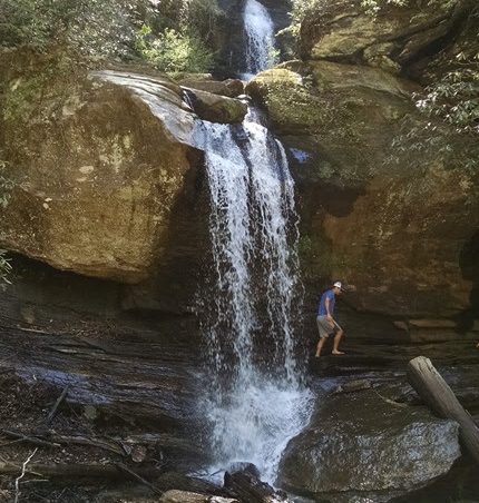 Person walking near a two-tiered waterfall in a forested area.