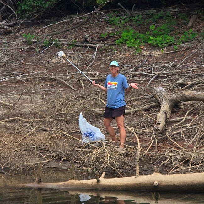 Person cleaning riverbank with grabber and trash bag.