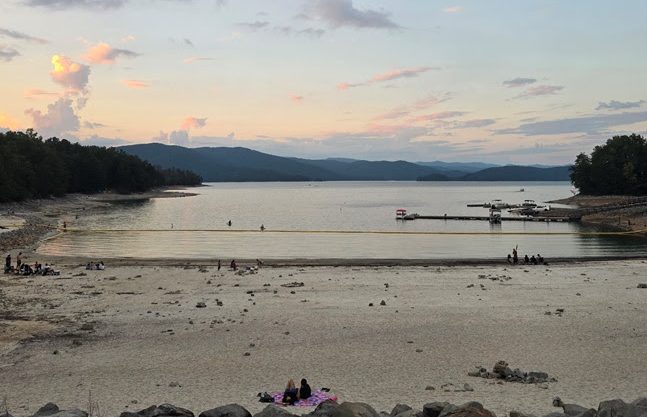 People on a sandy beach near a calm lake with a pier at sunset.