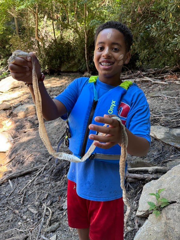 Boy in life jacket holds snake skin near a rocky forest area.