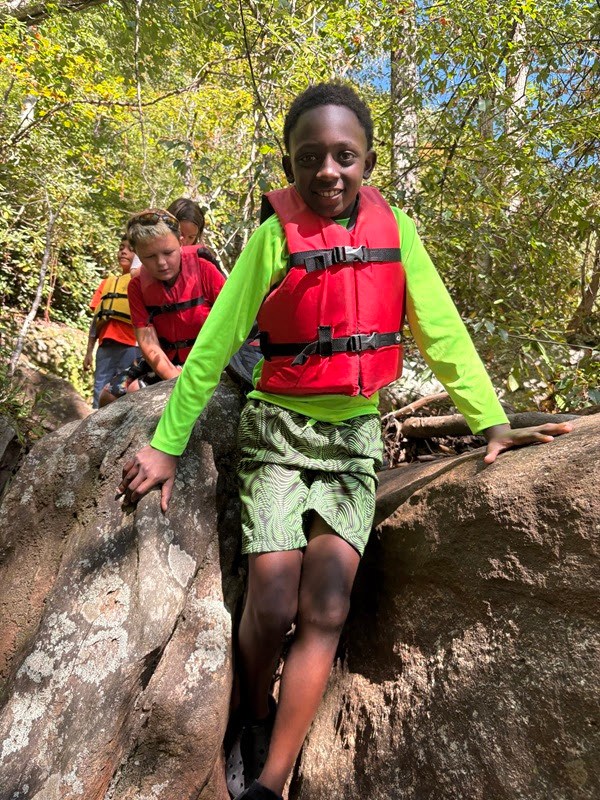 Children wearing life vests climb over rocks in a forest area.