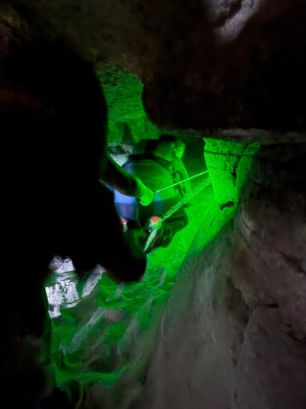 Person in dark cave illuminated by green light, holding a rope.